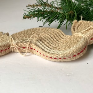 May include: A beige woven fabric ornament shaped like a candy wrapper, featuring a textured surface with a single horizontal red stitched line running through the center. The ends are gathered and tied with thin white string, creating a knotted effect.