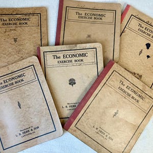 May include: A collection of five vintage tan exercise books titled The Economic Exercise Book. Each notebook features a black border, a central decorative icon, and the manufacturer text L. B. Herr & Son, Lancaster, PA printed on the front cover.