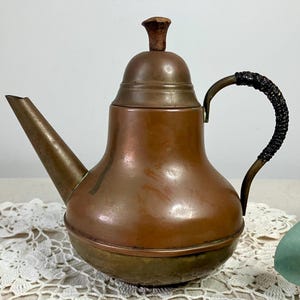 May include: Vintage copper teapot with a dark brown handle and a wooden knob on the lid. The teapot has a spout and a rounded body. The handle is wrapped in a dark material. The teapot is sitting on a white lace doily.