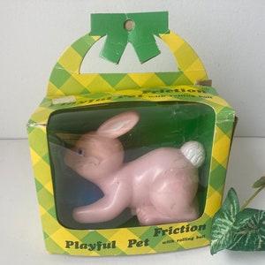 May include: A vintage pink plastic toy bunny inside a cardboard box with a green and yellow checkered pattern. The box reads "Playful Pet" and "Friction with rolling ball". The bunny has blue eyes and a white fluffy tail.