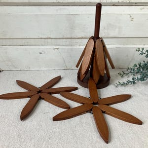 May include: A set of three brown wooden trivets, each featuring six articulated, petal-shaped slats that expand into a star shape. One trivet is stored vertically on a central wooden stand, while two others are laid flat in a star configuration.