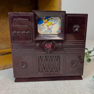 May include: A vintage maroon plastic toy television with a small screen displaying a cartoon image. The toy has a retro design with buttons, a dial, and decorative elements. The television is a collectible item.
