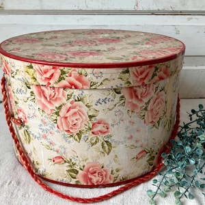 May include: A round, vintage-style hat box with a floral rose pattern in pink and cream. The box has a red trim and a red rope handle. The background is a white, distressed wooden surface.