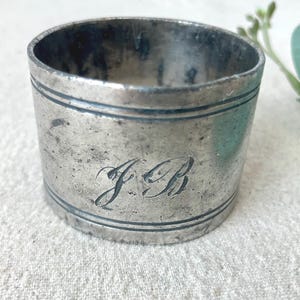 May include: A silver-colored napkin ring with engraved initials "J B". The ring has a cylindrical shape with two engraved lines. The metal has a worn, vintage appearance. The background includes a white cloth and green foliage.