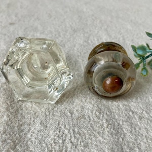 May include: Two vintage glass cabinet knobs. One is a clear, faceted hexagon shape. The other is a round knob with a layered, clear glass design and a brown center. Both are likely antique or vintage cabinet hardware.