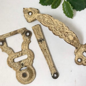 May include: An antique cream-colored door handle set with ornate floral detailing. The set includes a long handle, a keyhole plate, and a smaller piece, all showing signs of wear.