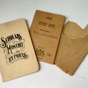 May include: Three vintage paper documents. The first is a booklet titled "Scholars Monthly Reports" in ornate black lettering. The second is a "School Report Book" with a name and the year 1904. The third is a torn paper envelope.