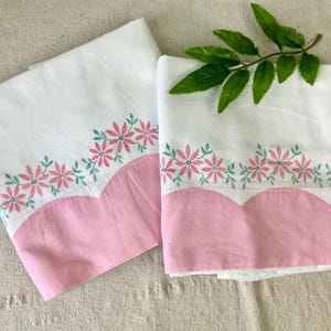 May include: Two white pillowcases featuring a scalloped pink bottom edge and a horizontal row of embroidered pink flowers with green stems and leaves. The fabric is smooth and clean, with a small green leafy branch resting on the right pillowcase.