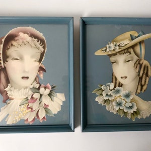 May include: Set of two framed vintage art prints. Each print features a woman's portrait with a hat and floral accents. The frames are a muted blue color, and the artwork has a soft, pastel color palette.