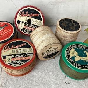 May include: A collection of vintage fishing line spools. The spools are round with various colors and labels, including "Shakespeare Wexford Wonder Line" and "Surf Comber Bait Casting Line." The spools are filled with tan, green, and brown line.