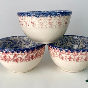 May include: Three off-white ceramic bowls with a speckled pattern in shades of blue and red. The rims are a solid dark blue. These bowls are suitable for serving soup, cereal, or other dishes.