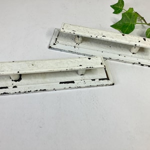 May include: Two rectangular, distressed white metal drawer pulls with a raised handle and a black border. The pulls have a vintage, rustic appearance, with chipped paint and visible wear. Green leaves are in the background.