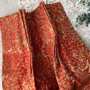 May include: Close-up of a folded fabric with a floral pattern. The fabric features a red and gold brocade design with detailed botanical motifs. The texture appears rich and luxurious, suggesting a decorative textile.