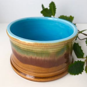 May include: A handcrafted ceramic bowl with a vibrant color scheme. The interior is a bright blue, transitioning to bands of green, brown, and tan on the exterior. The bowl has a rounded base and a wide opening.