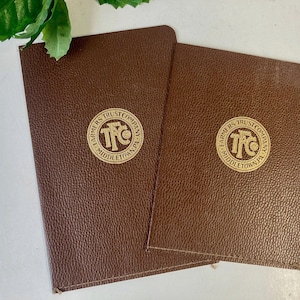 May include: Two brown textured notebooks with rounded corners. Each notebook features a circular gold logo in the center with the text Farmers Trust Company Middletown, PA and the letters FTC. The covers have a pebbled, leather-like appearance.