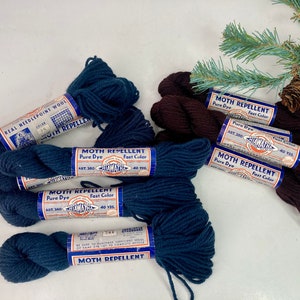 May include: Bundles of navy blue and brown yarn with labels reading "Moth Repellent" and "Real Needlepoint Wool." The yarn is arranged on a white surface with a sprig of greenery in the background. Each bundle contains 40 yards.