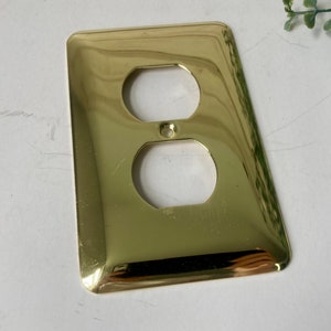May include: A shiny, gold-colored electrical outlet cover with two openings and a central screw hole. The cover has a rectangular shape with slightly curved edges. The surface reflects light, giving it a polished appearance. The item is likely for home improvement or electrical projects.