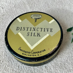 May include: A round, vintage tin with a black and gold rim and a gold and cream-colored top. The text "DISTINCTIVE SILK" is printed in black, with "Underwood Corporation, Burlington, N.J." below.