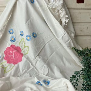 May include: White fabric with embroidered floral design. A large pink rose is the focal point, surrounded by blue and yellow daisy-like flowers and green leaves. The fabric is draped, with a fringe detail and greenery in the background.