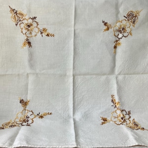 May include: White linen tablecloth with embroidered floral corner designs in shades of brown and gold. The embroidery features detailed flower and leaf patterns, adding a touch of elegance to the fabric. The tablecloth is folded, showing the embroidered corners.