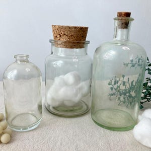 May include: Three clear glass bottles of varying sizes. The bottles have cork stoppers. One bottle contains cotton balls. The bottles are arranged on a light-colored surface with a sprig of greenery.