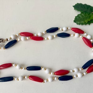 May include: A necklace featuring a patriotic color scheme. The necklace has alternating red, white, and navy blue beads. The beads are connected by small gold-colored beads and a gold-colored clasp.