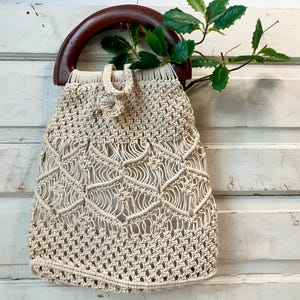 May include: Cream-colored macrame handbag with a dark brown wooden handle. The bag features a geometric pattern and a loop closure. Green leaves are placed near the top of the bag, adding a natural touch. The bag is displayed against a white, weathered background.