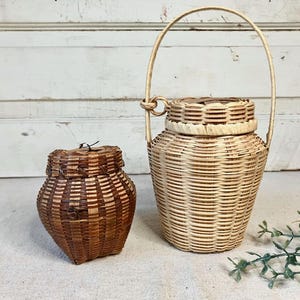 May include: Two woven baskets with lids. The smaller basket is dark brown with a rounded shape. The larger basket is light beige, features a tall arched handle attached with a ring, and has a woven lid. Both baskets have a textured finish.