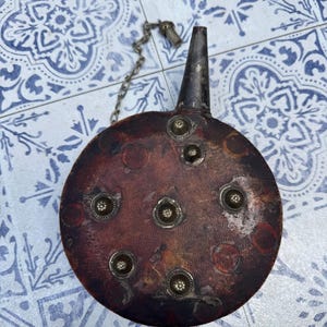 Könnte beinhalten: Eine runde, antike Lederflasche mit dunkelbraunem Finish. Die Flasche hat dekorative Metallnieten und einen Metallausguss mit Kette und Verschluss. Die Flasche wird wahrscheinlich zum Tragen von Flüssigkeiten verwendet.