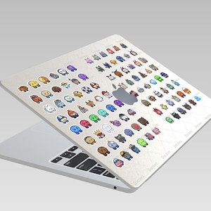 May include: A silver laptop with a white skin featuring a grid pattern of numerous small, colorful cartoon character stickers. The stickers depict various creatures, objects, and figures in diverse colors, arranged in rows across the laptop lid.