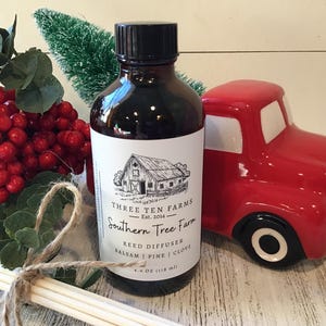 May include: A dark brown glass bottle with a black cap and a white label featuring a barn illustration. The label reads "Southern Tree Farm" and lists "Balsam, Pine, Clove". A red ceramic truck and reed diffuser sticks are also visible.