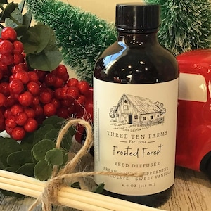 May include: A dark brown glass bottle with a black cap and a white label featuring a barn illustration. The label reads "Frosted Forest" and "Reed Diffuser." The bottle is next to a red truck and red berries.