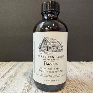 May include: A dark glass bottle of Three Ten Farms Plantain Organic Infused Oil. The label features a barn illustration and text including "Plantain (Plantago major)" and "4.0 OZ (118 ml)". The bottle has a black cap.