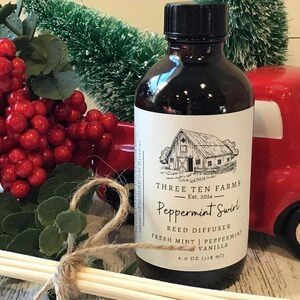 May include: A dark brown glass bottle with a black cap and a white label featuring a barn illustration. The label reads "Peppermint Swirl" and "Reed Diffuser." The bottle is next to a red truck and red berries.
