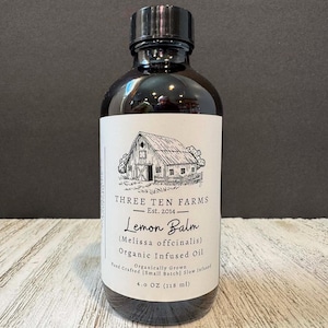 May include: A dark glass bottle with a black cap, labeled "THREE TEN FARMS" and "Lemon Balm (Melissa officinalis) Organic Infused Oil." The label features a barn illustration and states the product contains 4.0 OZ (118 ml).
