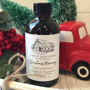 May include: A dark brown glass bottle of reed diffuser with a black cap, labeled "Christmas Morning" by Three Ten Farms. The label features a barn illustration. A red truck and red berries are in the background. The bottle contains 4.0 oz (118 ml).