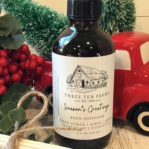 May include: A dark brown reed diffuser bottle with a black cap, featuring a label with a barn illustration and the text "Season's Greetings." The bottle is 4.0 oz (118 ml). The image includes a red truck and decorative berries.