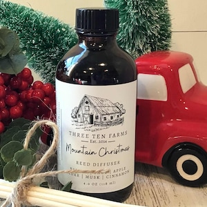 May include: A brown glass bottle with a black cap and a white label featuring a barn illustration. The label reads "Mountain Christmas Reed Diffuser." A red truck and decorative berries are in the background. The bottle contains 4.0 oz (118 ml).