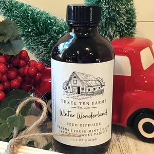 May include: A dark brown glass bottle with a black cap, labeled "Winter Wonderland Reed Diffuser." The label features a barn illustration and scent notes. A red truck and decorative berries are in the background. The bottle contains 4.0 oz (118 ml).