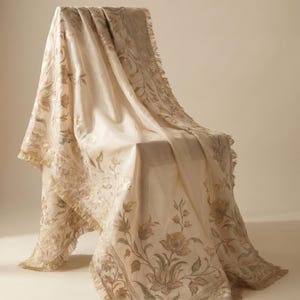 May include: A cream-colored throw blanket featuring an intricate floral pattern in muted gold and sage green tones. The fabric is draped over a chair and is finished with a delicate fringe trim along the edges, showcasing detailed botanical embroidery.