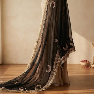 May include: A black sheer shawl with gold and red embroidered crescent moons and stars. The shawl has a gold fringe trim along the edges. The fabric drapes over a beige garment, creating a flowing effect.