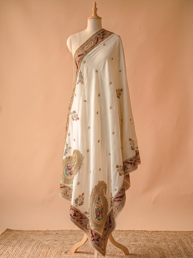 May include: An off-white shawl draped on a mannequin, featuring intricate gold embroidery, deep red velvet borders, and colorful floral paisley motifs. The edges are finished with delicate pearl-like beadwork and gold thread detailing.