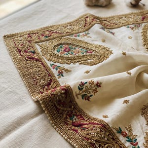 May include: An off-white fabric textile featuring a wide border in deep burgundy with intricate gold metallic embroidery, beadwork, and sequins. The center displays paisley motifs and small floral sprigs in green, pink, and orange thread.
