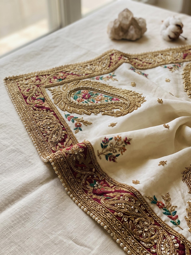May include: An off-white fabric textile featuring a wide border in deep burgundy with intricate gold metallic embroidery, beadwork, and sequins. The center displays paisley motifs and small floral sprigs in green, pink, and orange thread.