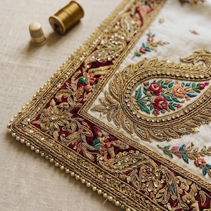 May include: A white fabric panel featuring intricate gold metallic embroidery, a deep maroon border, and colorful floral motifs. The edge is finished with small white beads. A gold thimble and a spool of gold thread are placed nearby.