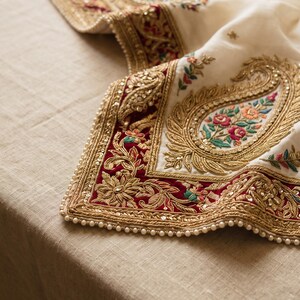 May include: An off-white fabric runner featuring intricate gold metallic embroidery, deep red velvet borders, and colorful floral motifs. The edges are adorned with small white beads, creating a textured and ornate decorative textile piece.