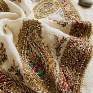 May include: An off-white fabric featuring intricate gold embroidery in a large paisley pattern. The design includes colorful floral accents in red, green, and blue, with metallic sequins and beads. A deep maroon border with gold embellishments frames the edge.