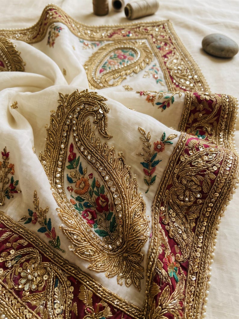 May include: An off-white fabric featuring intricate gold embroidery in a large paisley pattern. The design includes colorful floral accents in red, green, and blue, with metallic sequins and beads. A deep maroon border with gold embellishments frames the edge.