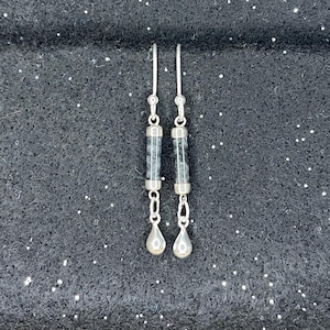 May include: A pair of dangle earrings with a silver finish. Each earring features a cylindrical, clear bead with a speckled, dark gray pattern, suspended from a silver hook. Below the bead hangs a silver teardrop-shaped charm.