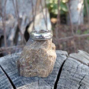May include: A silver ring with a detailed floral pattern rests on a rough, brown stone. The ring is displayed on a weathered wooden surface, with a blurred natural background.